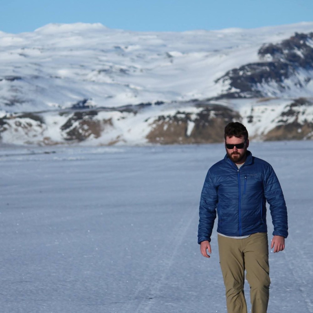 Man in blue jacket walking on a snowy landscape with mountains in the background.