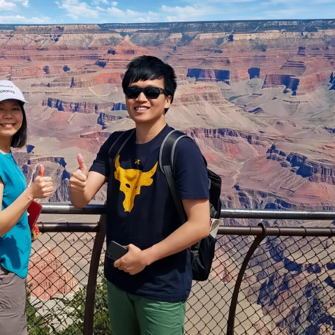 Two people smile and give thumbs up at Grand Canyon railing.