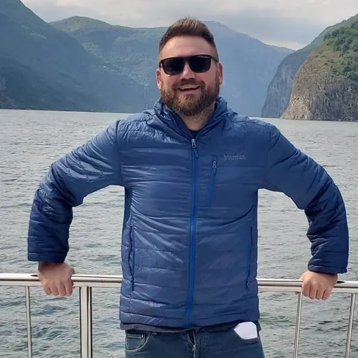 Person in blue jacket and sunglasses standing by railing with mountains and water in the background.